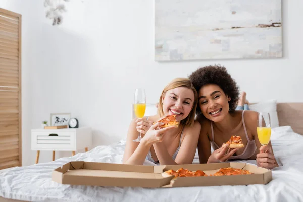Joyful multiethnic women looking at camera while lying on bed with delicious pizza and champagne glasses with cocktails — Stock Photo