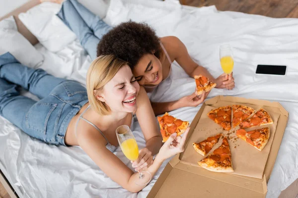 High angle view of excited multiethnic women holding delicious pizza and cocktails while lying on bed at home — Stock Photo