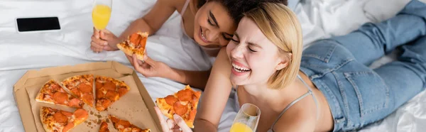 Високий кут зору веселих міжрасових жінок з коктейлем і смачною піцою, що лежить на ліжку вдома, банер — стокове фото