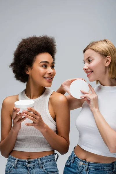 Mulheres muito inter-raciais em tops brancos segurando recipientes com creme cosmético e sorrindo um para o outro isolado em cinza — Fotografia de Stock