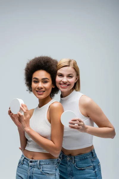 Досить багатоетнічні моделі в білих баках позують з контейнерами косметичного крему ізольовані на сірому — стокове фото