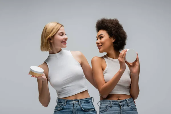 Молоді багатоетнічні жінки тримають крем для тіла і посміхаються один одному ізольовані на сірому — стокове фото