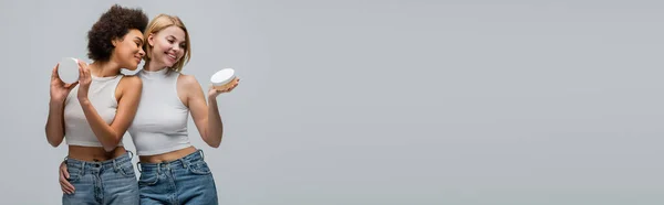 Жизнерадостная блондинка-модель, обнимающая африканскую американку, держа косметический крем изолированный на сером, баннер — стоковое фото