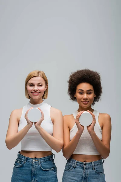 Вид спереду молодих міжрасових жінок в білих баках і тримає банки з косметичним кремом ізольовані на сірому — стокове фото
