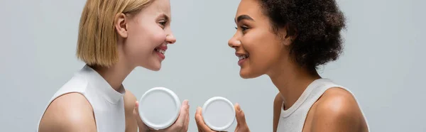 Vista lateral de mujeres rubias y morenas interracial con crema corporal sonriéndose entre sí aisladas en gris, pancarta — Stock Photo