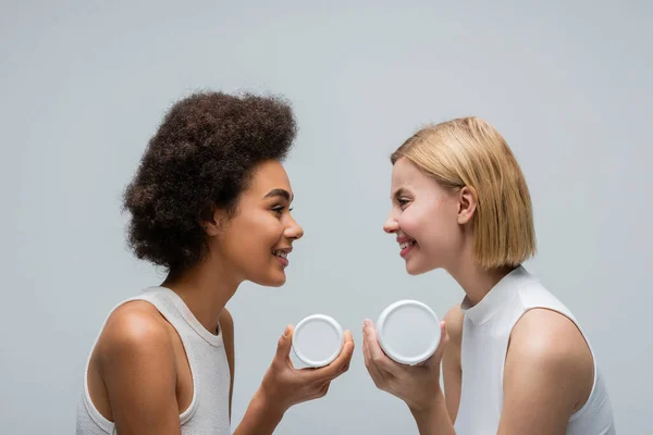 Vista lateral de mulheres loira e morena multiétnica segurando creme cosmético e sorrindo um para o outro isolado em cinza — Fotografia de Stock