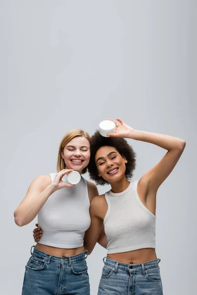 African american model hugging blonde woman posing with jar of cosmetic cream isolated on grey - foto de stock