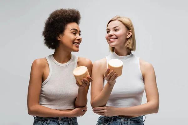 Pleased multiethnic models in white tank tops holding containers with body cream isolated on grey — Stock Photo