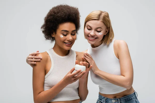 Interracial blonde und brünette Frauen lächeln in der Nähe Glas Gesichtscreme isoliert auf grau — Stockfoto