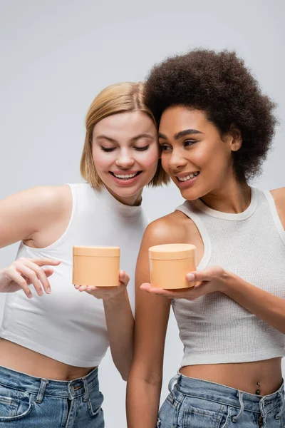 Молода афроамериканка тримає банки з косметичним кремом біля блондинки, вказуючи пальцем ізольовано на сірому — стокове фото