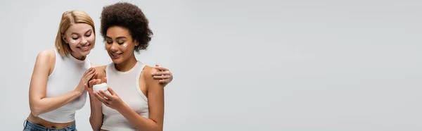 Усміхнена блондинка модель торкається плечей афроамериканки тримає крем для обличчя ізольовано на сірому, банер — стокове фото