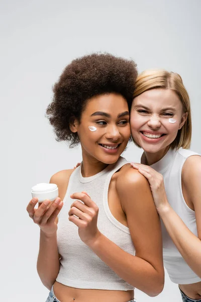 Cheerful blonde woman looking at camera near african american model posing with face cream isolated on grey — Stock Photo