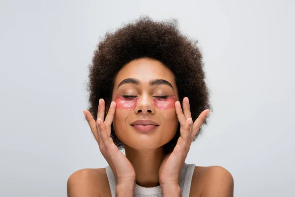 Mujer afroamericana bonita con los ojos cerrados aplicando parches hidrogel aislados en gris - foto de stock