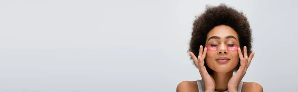 Brunette african american woman with closed eyes applying lifting eye patches isolated on grey, banner — Stock Photo