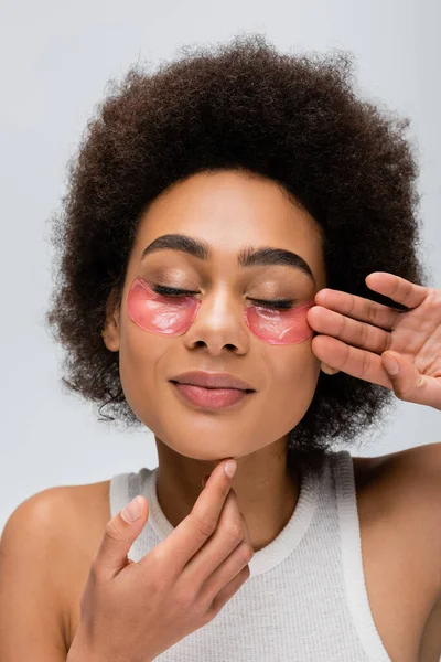 Young and positive african american woman touching face with hydrogel eye patches isolated on grey — Stock Photo