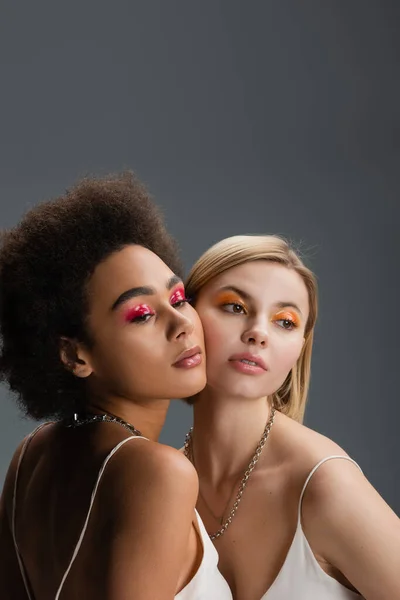 Sensual interracial women with bright and glossy eye shadows posing isolated on grey — Stock Photo