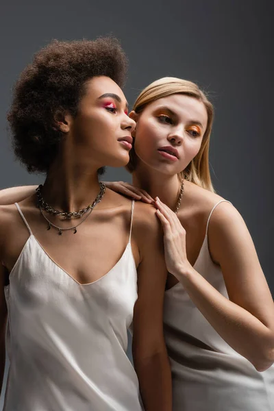 Sensual interracial women with colorful makeup posing in strap dresses and silver necklaces isolated on grey — Stock Photo