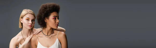 Young african american model in white strap dress looking away near blonde woman with bright makeup isolated on grey, banner — Stock Photo