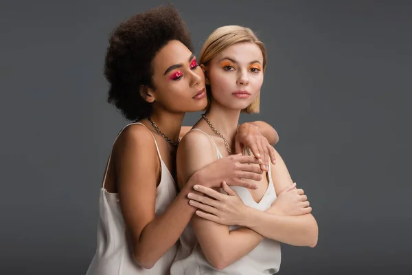 Young multiethnic models with colorful makeup posing in white strap dresses isolated on grey — Stock Photo
