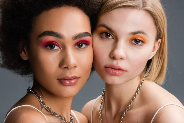 Portrait of young multiethnic women with bright and glossy eye shadows and silver necklaces isolated on grey — Stock Photo