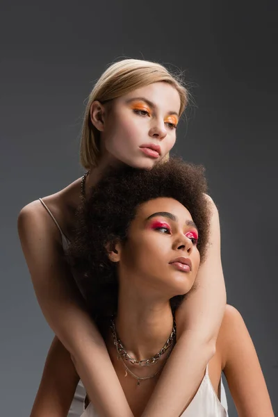 Young blonde model with bright visage embracing african american woman looking away isolated on grey — Stock Photo