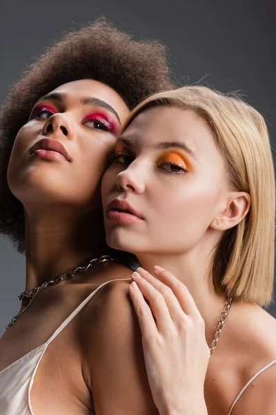 Blonde woman with bright makeup looking at camera near african american model isolated on grey — Stock Photo