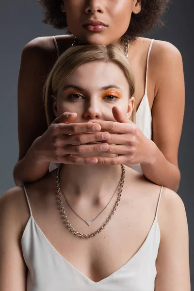 African american woman covering mouth of blonde model in white strap dress and silver necklaces isolated on grey — Stock Photo