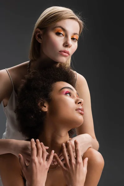 Blonde model with orange eye shadows embracing brunette african american woman while looking at camera isolated on grey — Stock Photo