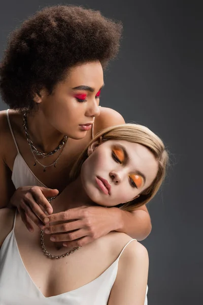 Young african model in bright makeup and silver necklaces embracing blonde woman isolated on grey — Stock Photo