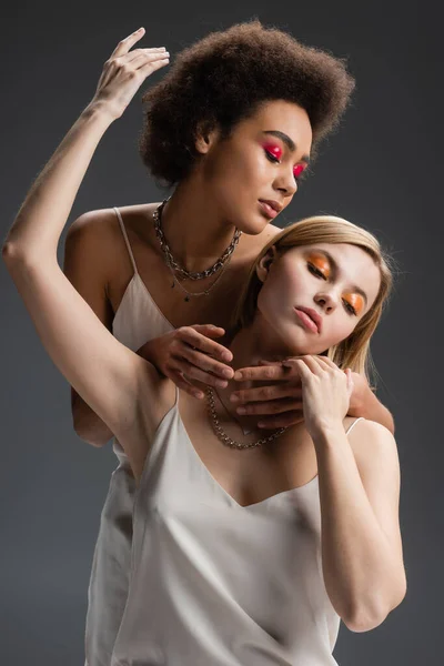 Sensual interracial models in white strap dresses and colorful makeup posing isolated on grey — Stock Photo