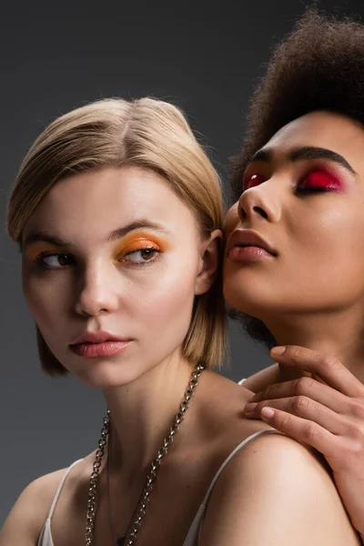Blonde woman with orange eye shadows looking away near sensual african american model posing with closed eyes isolated on grey — Stock Photo