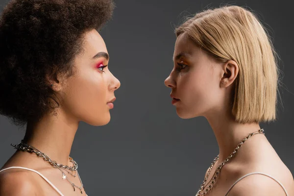 Side view of blonde and brunette interracial models in bright visage and silver necklaces isolated on grey — Stock Photo