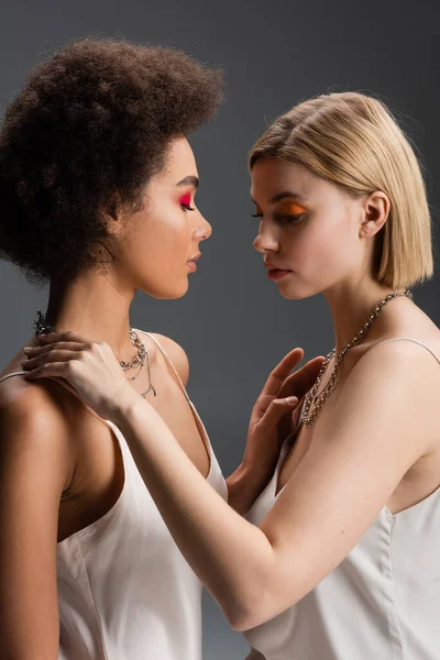 Side view of young multiethnic women in strap dresses and silver necklaces touching each other isolated on grey — Stock Photo