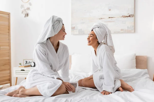 Full length of barefoot interracial women in white robes and towels sitting on bed and smiling at each other - foto de stock