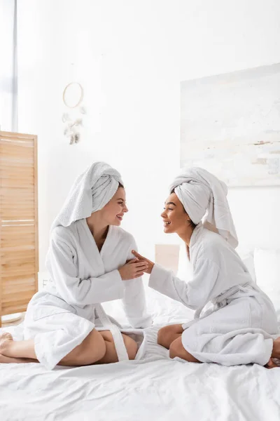 Joyful interracial women in white bathrobes and towels looking at each other while sitting on bed - foto de stock
