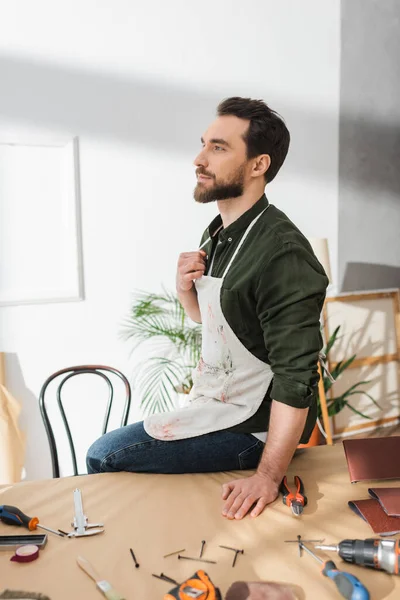 Bearded carpenter in apron sitting near tools on table in workshop — Stock Photo