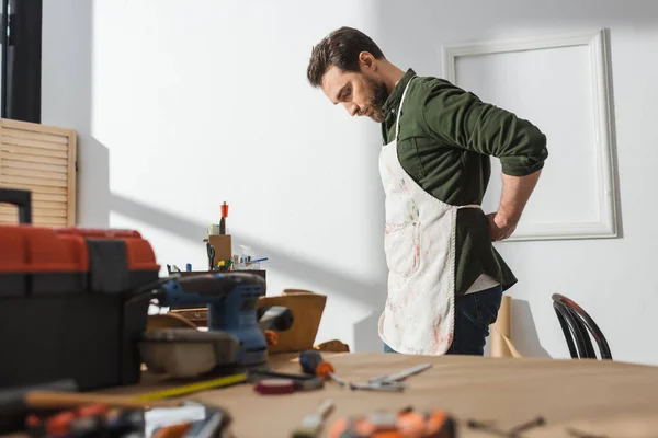 Side view of bearded carpenter wearing apron near blurred tools in workshop — Stockfoto