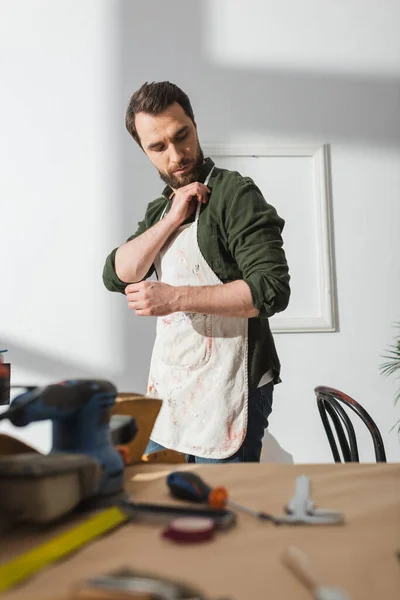 Bärtiger Handwerker mit Schürze nahe verschwommenem Werkzeug auf Tisch in Werkstatt — Stockfoto