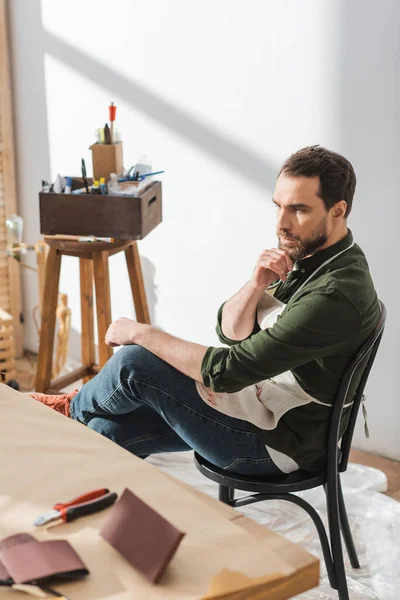 Artigiano pensieroso in grembiule seduto sulla sedia vicino al tavolo sfocato in officina — Foto stock