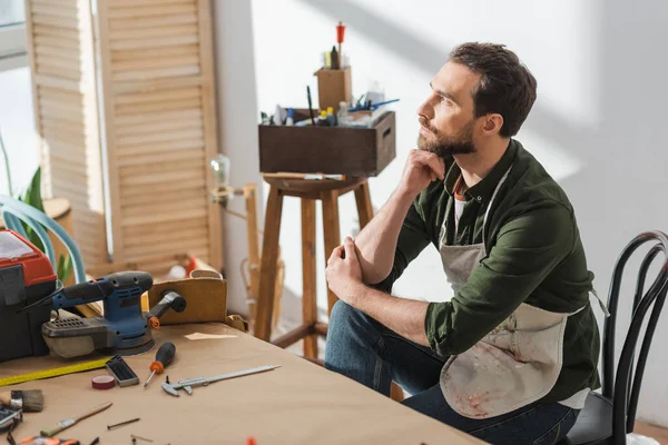 Thoughtful carpenter in apron sitting near tools on table in workshop — Stock Photo