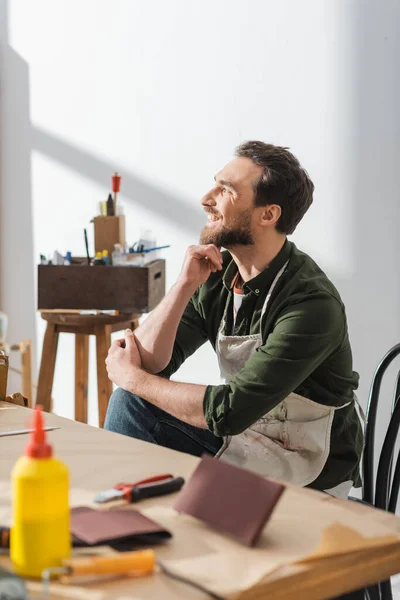Carpintero sonriente en delantal sucio sentado cerca de la mesa de trabajo en el taller - foto de stock