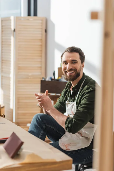 Smiling carpenter in apron looking at camera while sitting in workshop — Foto stock