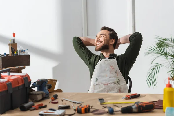 Artesãos alegres em avental sujo descansando perto de ferramentas borradas na mesa na oficina — Fotografia de Stock