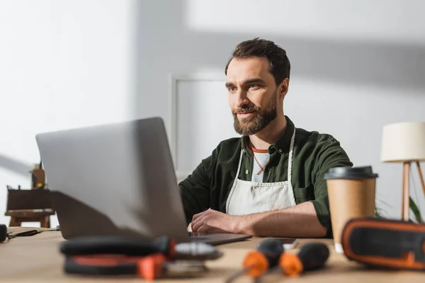 Bearded carpenter using laptop near blurred takeaway coffee and tools in workshop — Foto stock