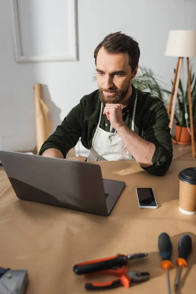 Artigiano in grembiule con computer portatile vicino strumenti e caffè per andare in officina — Foto stock