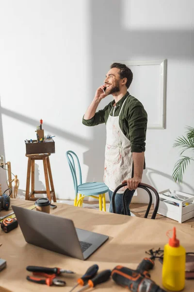 Positive craftsman talking on cellphone near tools and laptop on working table — Photo de stock