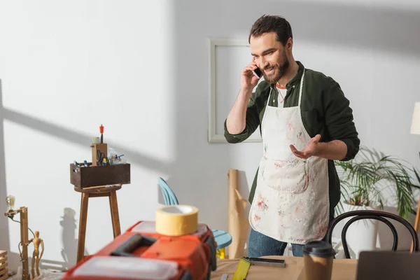 Smiling craftsman in apron talking on mobile phone near blurred toolbox in workshop — Stockfoto