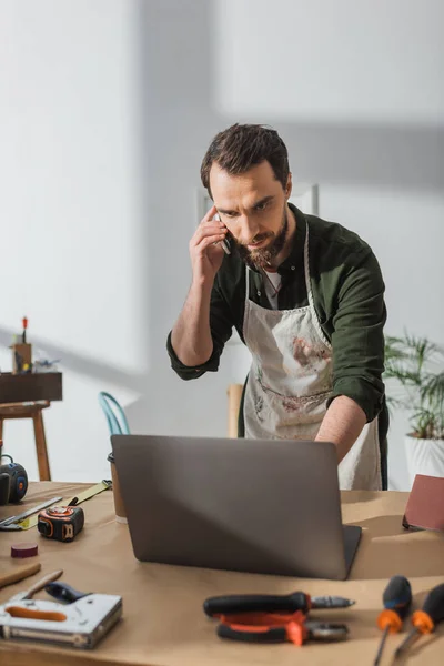 Bearded carpenter talking on smartphone and using laptop in workshop — Stock Photo