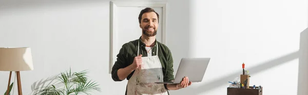 Cheerful carpenter in apron holding laptop and looking at camera in workshop, banner — Foto stock