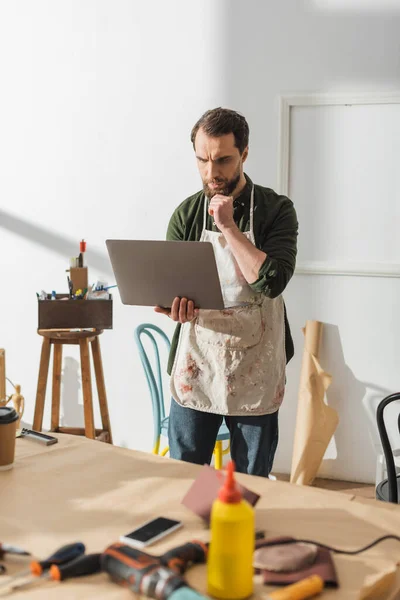 Pensive carpenter in apron using laptop in workshop — Stock Photo